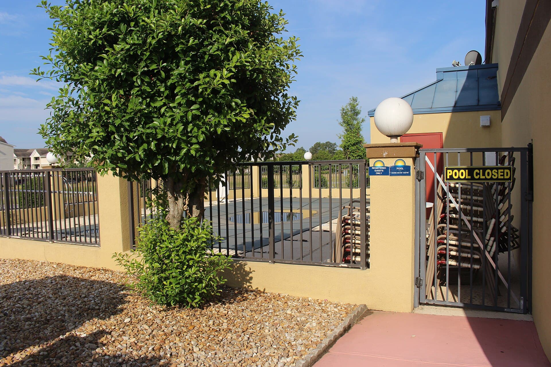 A 'Pool Closed' sign posted on a locked gate in front of a covered pool with stacked lounge chairs