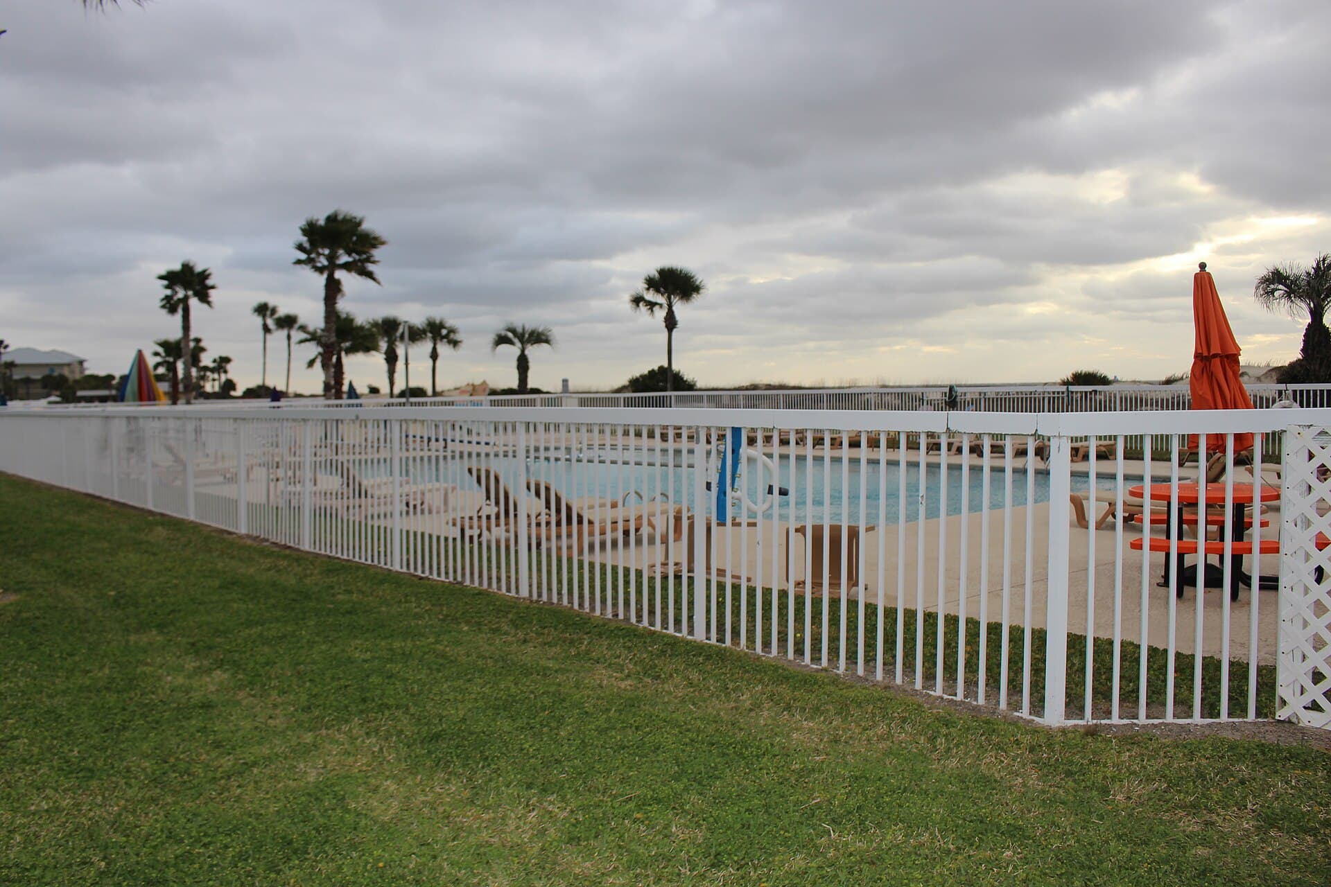 White vertical-bar safety fence enclosing an outdoor pool, with lounge chairs and palms behind the barrier