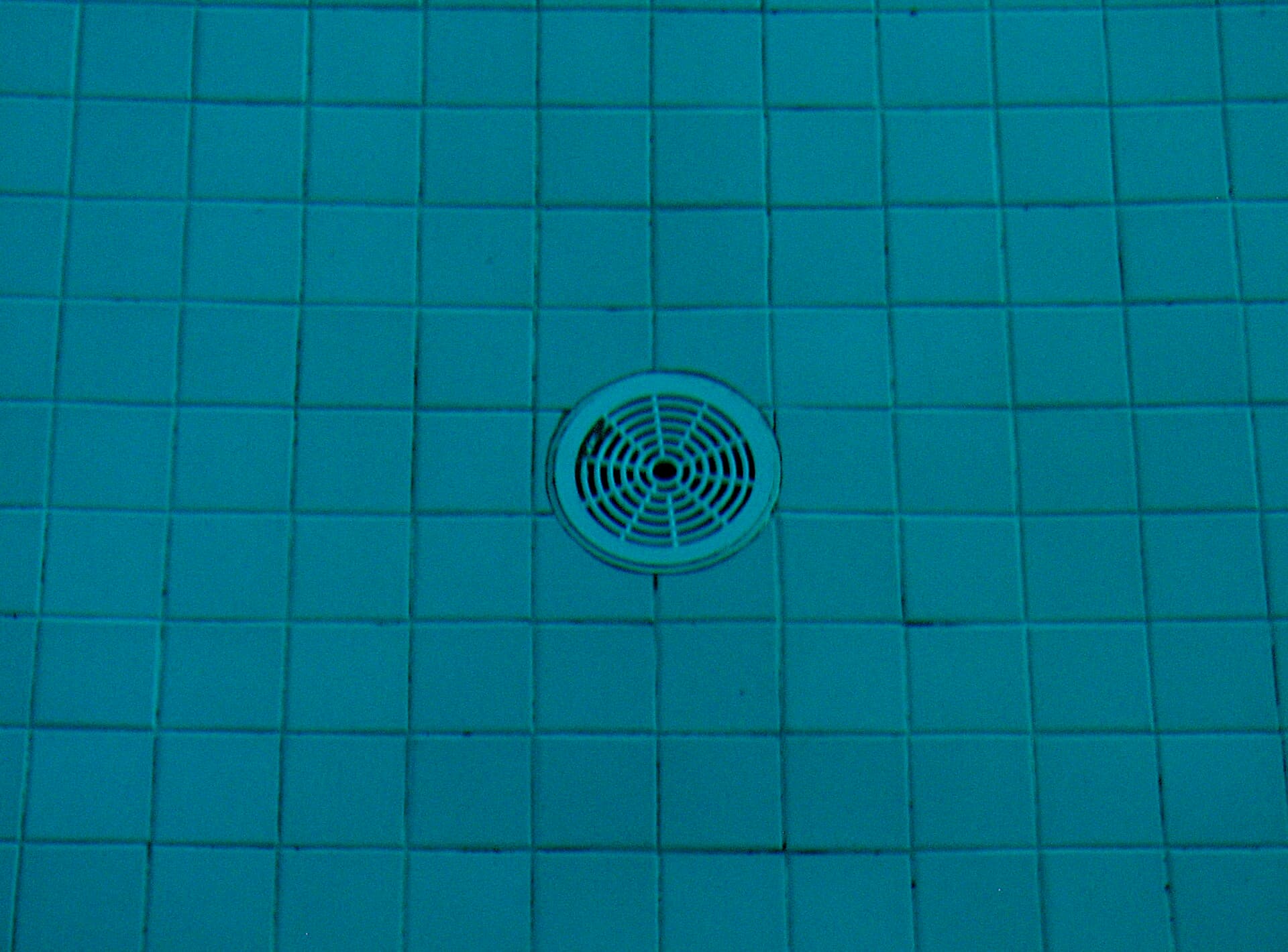 Round main drain cover on the tiled floor of a swimming pool, seen through clear blue water