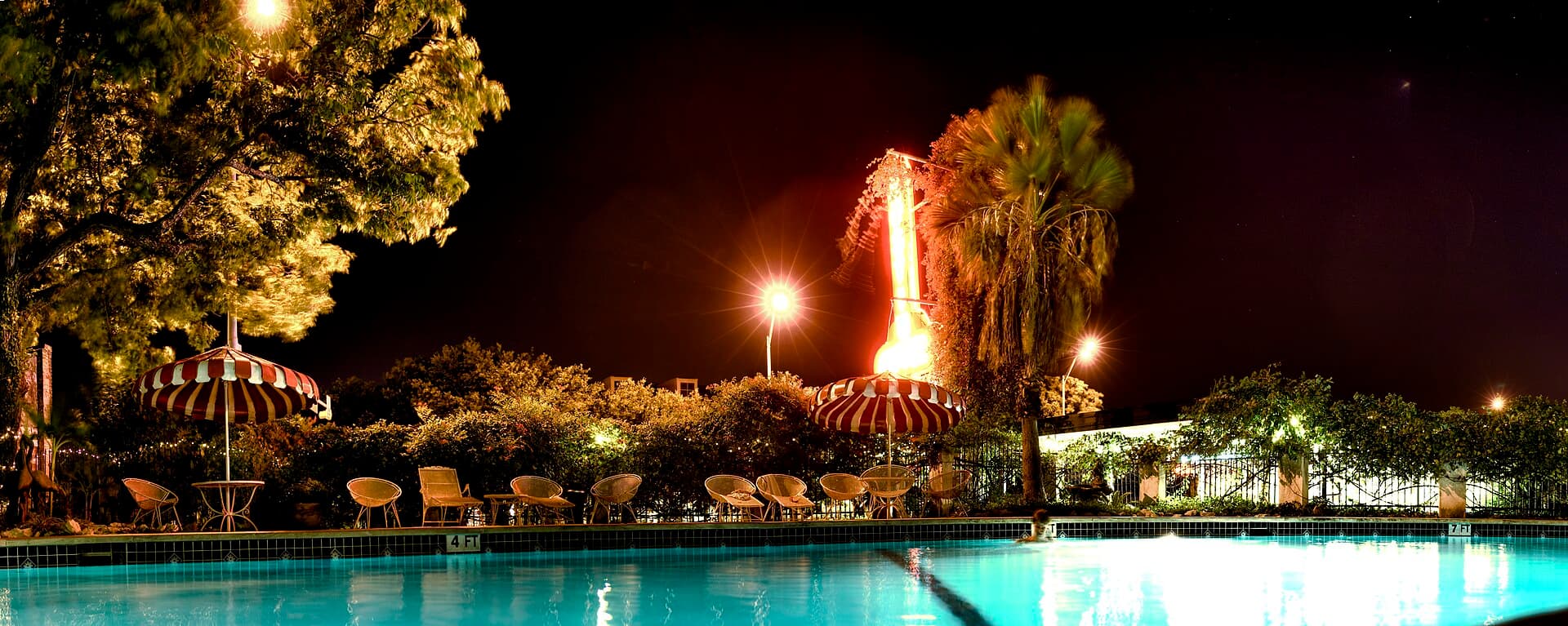 Motel swimming pool glowing at night with underwater lights, string lights, palm trees, and poolside umbrellas