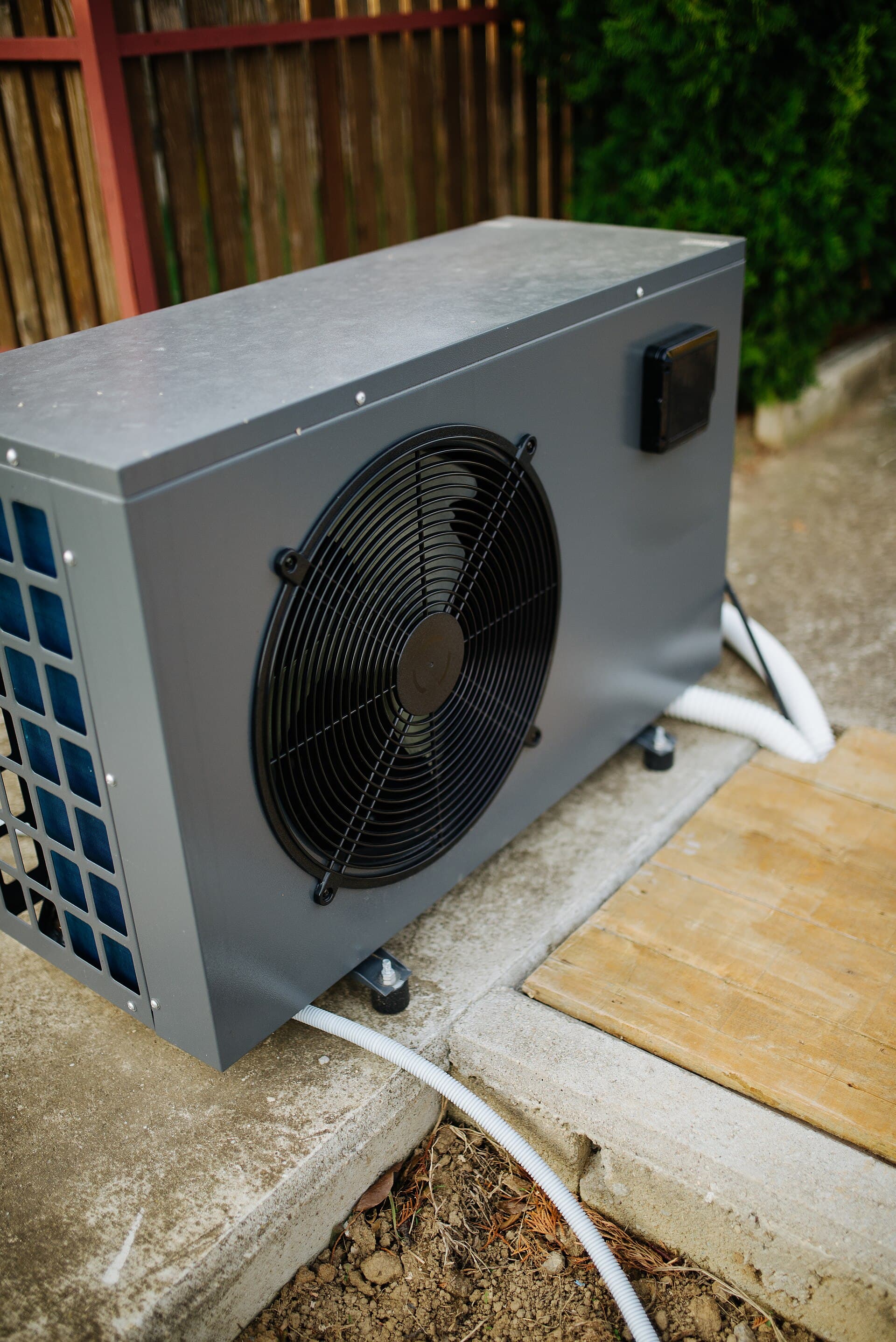 Pool heat pump heater unit sitting on a concrete pad beside a garden fence with plumbing lines running toward the pool