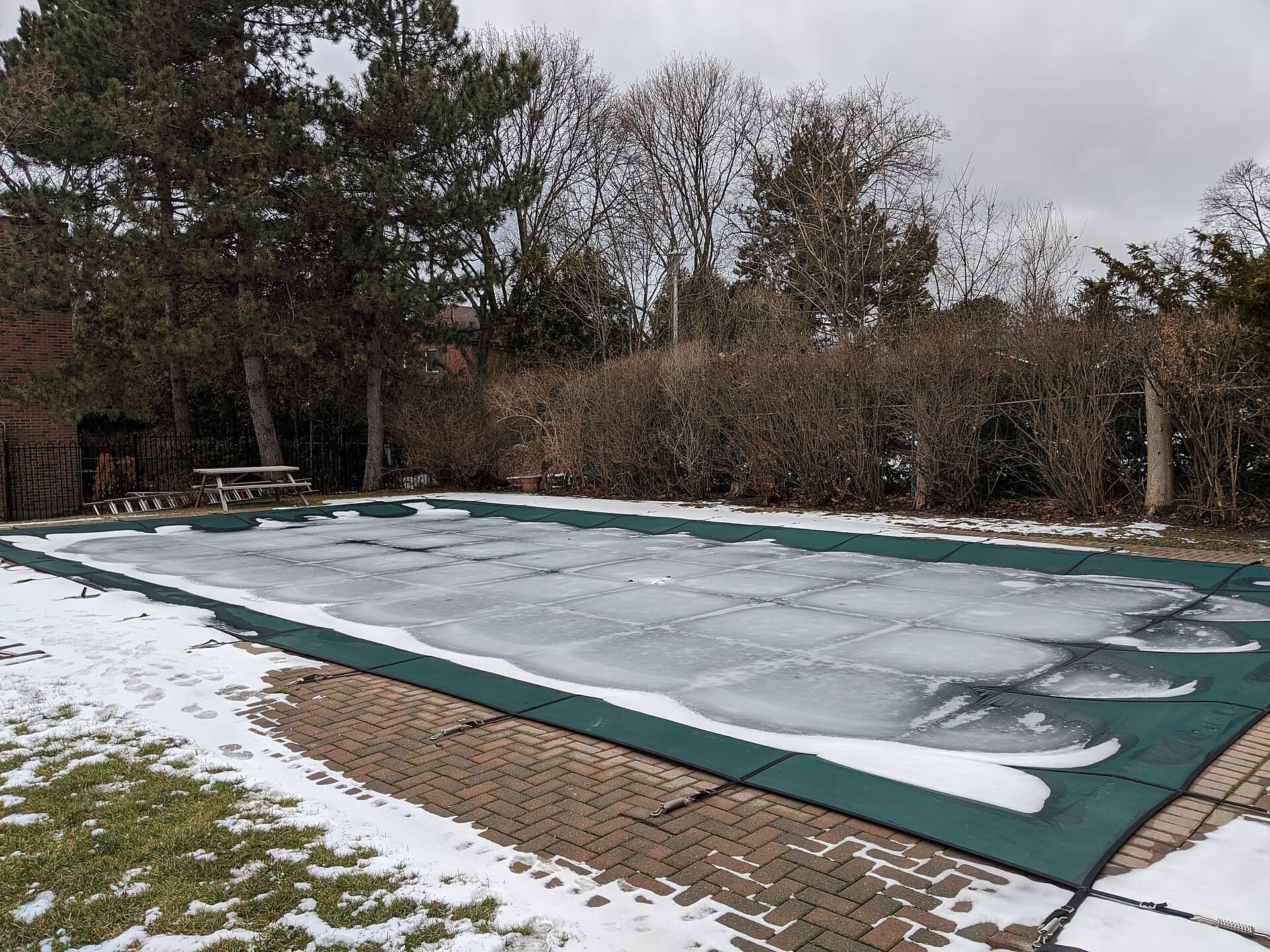 Backyard swimming pool closed for the winter under a green safety cover dusted with ice and snow