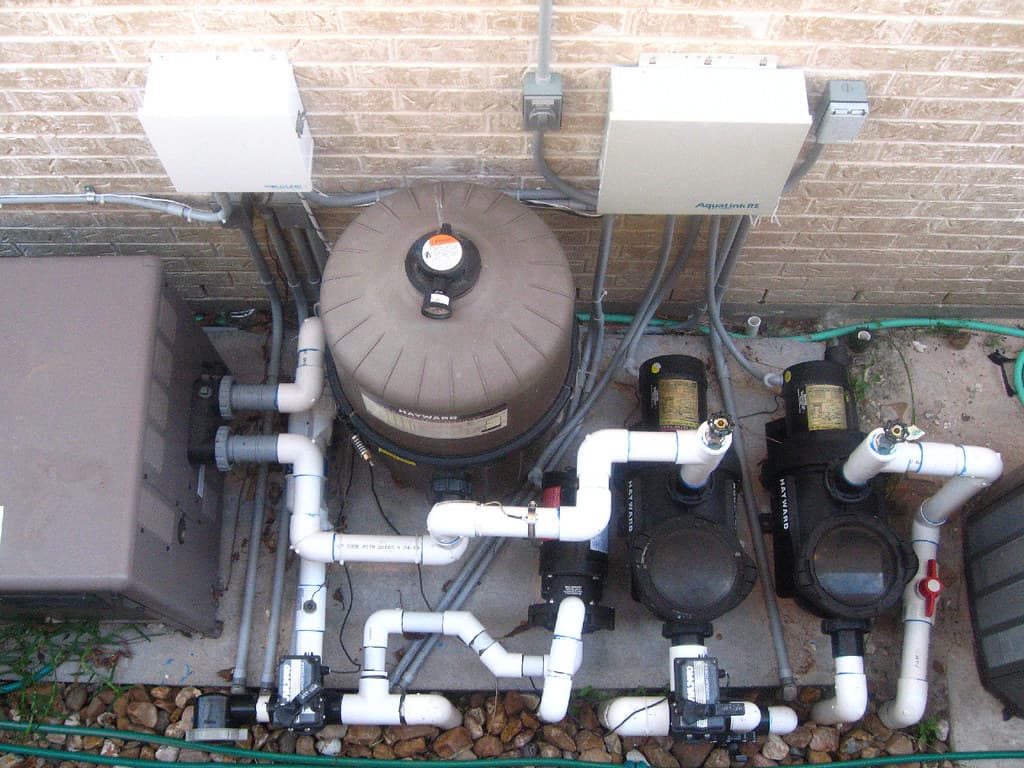 Residential pool equipment pad with pumps, cartridge filter, heater, control panels and PVC plumbing against a brick wall